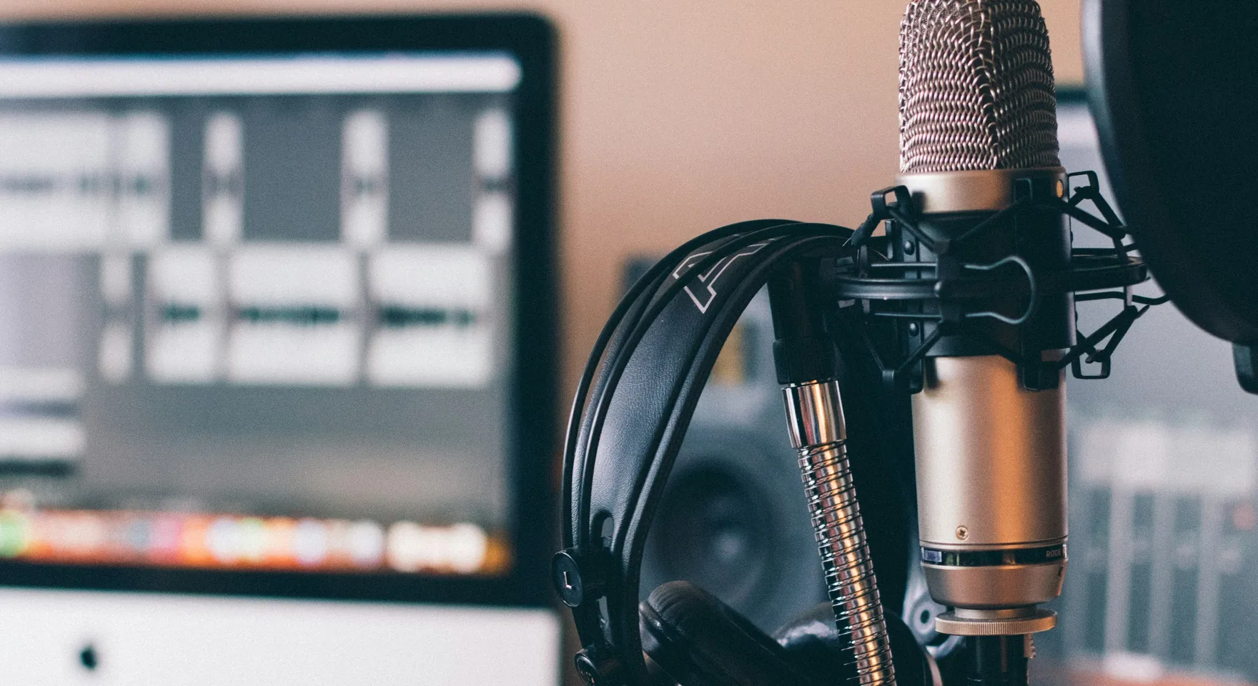 A condenser microphone on a shock mount behind a pop filter, with studio headphones beside it and an audio editing timeline on the screen behind.